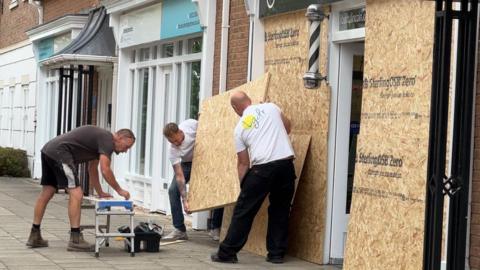 Three workmen installing a large plywood board onto windows in front of a shop