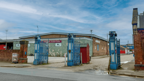 A large warehouse building behind the elaborate open iron gates painted blue