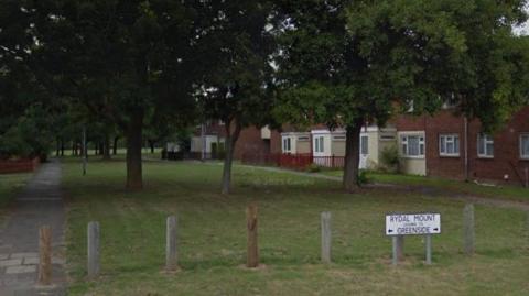 A road, with a row of wooden stumps, a street sign, and trees leading to a park. A row of houses are to the right.