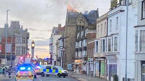 Police cars and van across a road with a fire engine in the distance - buildings are either side the roof of a church-like building on the right hand side is on fire.