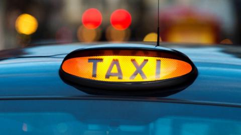 A stock image of the roof of a blue taxi with a "TAXI" light illuminated on it