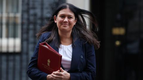 Lisa Nandy, a woman with long dark hair and wearing a white blouse and a navy coat. The entrance to Downing Street is in soft focus behind her and she's got a red folder tucked under arm.