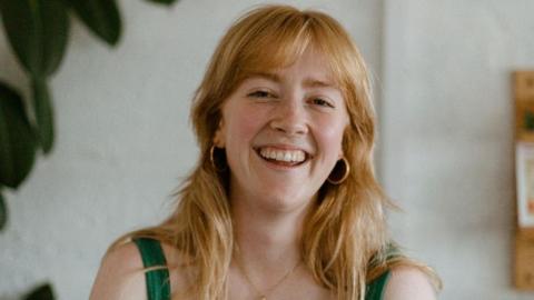 Ana Jaks has strawberry blonde hair and is wearing a green strappy top and gold earrings. She smiles at the camera.