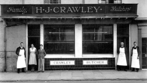 Black and white photo taken outside a butchers shopfront in the early 1900s. There are two windows with a sing above them that reads Family H.J Crawley Butchers. Underneath the windows there are tiles and the words family butcher. To the left of the widows and in front of a doorway are three people, one man dressed in a suit with an apron. A lady next to him wearing a pinafore dress and a younger man on her right. In the right of the photo there are two men wearing aprons over their day suits.