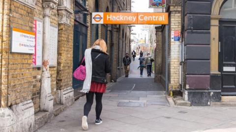 A woman in a black leather jacket, red skirt and black tights, with her back to the camera, walks into a passage - there is an orange and white sign that says Hackney Central above the passage.