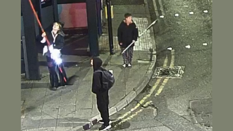 A CCTV image shows three people standing on a well-lit street corner at night. One person is holding a long red pole, another is holding a slightly shorter grey-coloured pole. A third person, at the forefront of the image, wears a dark top with the hood up, and is carrying a black backpack.