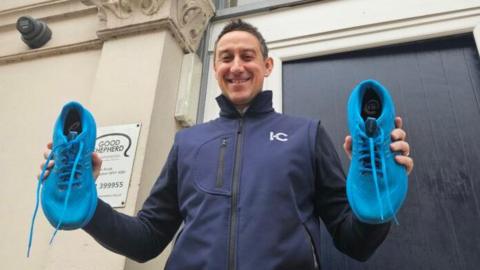 A man in a dark blue zipped jacket is holding bright blue running shoes and standing in front of a door