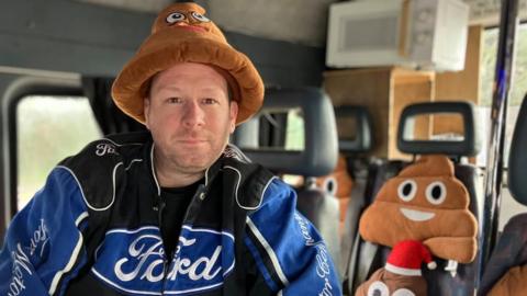 A man sitting in a minibus wearing a Ford jacket and a poo emoji hat. On the other seats in the bus are a number of poo emoji cushions, one wearing a Santa hat, and a microwave is on top of a wooden structure at the back of the bus.