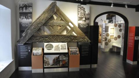 A room with a dark wood floor and exhibits on the wall, including a large stone roof-frame