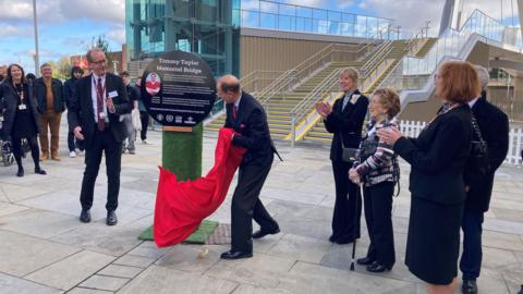 The Duke of Edinburgh pulls a sheet of the plaque dedicating the Tommy Taylor Memorial Bridge to the footballer. The crowd surrounding him is applauding