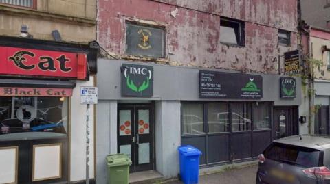 Exterior of a shabby building with black doors, grey windows and peeling brown paint on the first storey. Above the entrance is a sign saying "IMC" for Inverclyde Muslim Centre. It is next to a pub called the Black Cat.