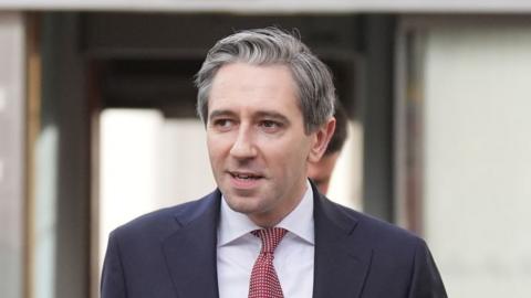 Simon Harris - a man with grey hair wearing a dark suit jacket, a white collared shirt and a red pattern tie walks outside a white stone building.