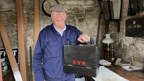 Phil Harris is holding a black wooden toolbox. It has red letters on it, reading "SVR". He is wearing a blue jacket, and a tweed cap. He is wearing glasses