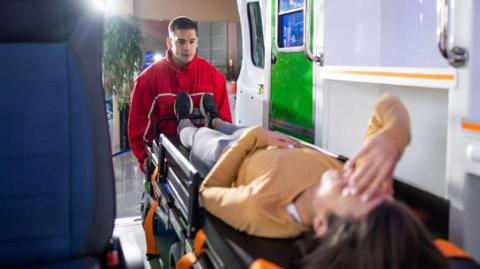 A paramedic transports a woman on a stretcher to an ambulance. She has her hand on her head. The ambulance doors are open.