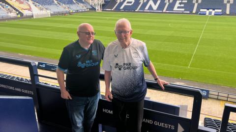 Peter and Terry Kilgariff are standing side by side in the stand at Deepdale Stadium - the home of Preston North End. They are both wearing Preston North End tops and the pitch can be seen behind them.