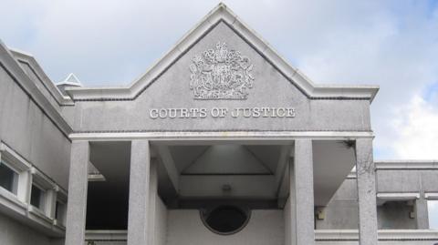 Truro Crown Court is pictured. It is made of pale grey granite and has four pillars and a steeple roof. There is an ornate heraldic shield on the front entrance and the words 'Courts of Justice'.