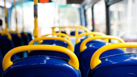 The inside of a bus, facing forward, showing the backs of blue seats with yellow handles on the top