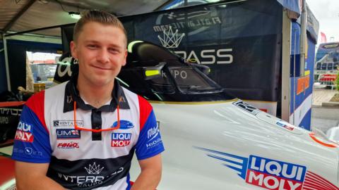 Powerboat racer Ben Jelf wearing a red, white and blue top while smiling at the camera. He is sat on the right side of his boat, which is on land