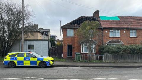A police car outside a brick terraced house. There is a gap in the roof and rafters are exposed. 