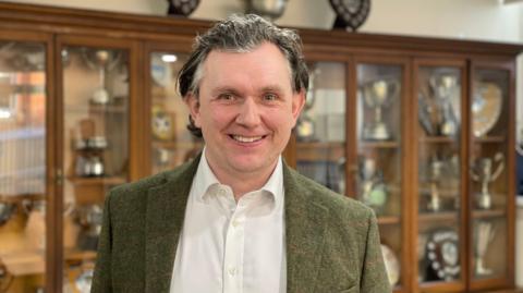 A man with long grey hair is wearing a white shirt and a green suit jacket. He is standing in front of cabinets with brown wood and glass doors.