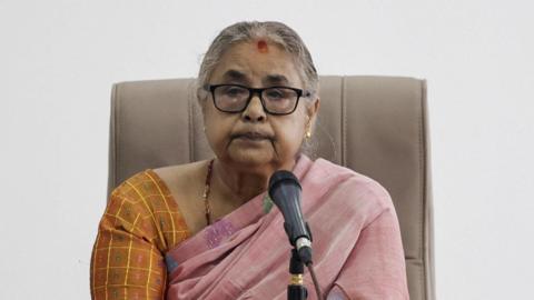 Interim Prime Minister Sushila Karki sits in a beige chair in front of a microphone, wearing an orange an pink outfit. She is wearing black framed glasses.