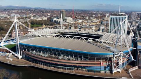 What is now the The Principality Stadium, viewed from the opposite side of the River Taff . Its four white-painted masts which hold up its retractable roof are visible. It's set against a blue sky and calm river and the city of Cardiff can be seen spreading out in the background behind it