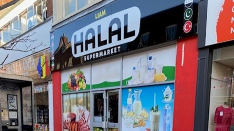 A shop with a black sign above the door that reads Liam HALAL Supermarket. The shop also has images of fruit and glasses of milk on the exterior of the building. 