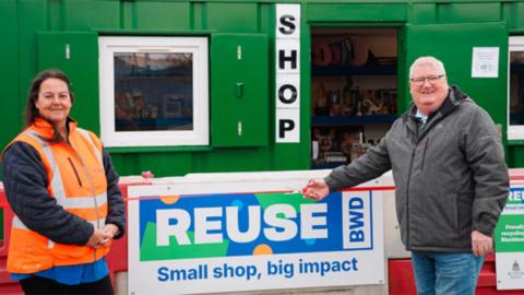 Victoria Crabtree and councillor Jim Smith outside the shop, which is a large green shipping container with windows. Victoris is standing to the left of a sign that says 'reuse small shop, big impact'. She is wearing an orange high vis vest and is smiling. Jim stands at the other side in a grey coat and jeans pointing at the sign
