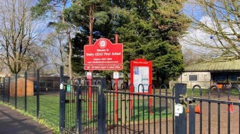 The grounds of Trinity First School. There is a black gate in the foreground. Two red signs are in the centre of the image. There is a building and a large tree in the background. There is a shed and other buildings to the left of the image.