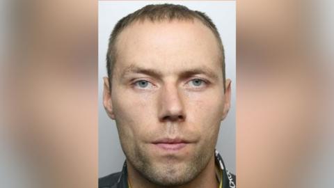 The mugshot of a man who looks at the camera. He has blue eyes and blond hair.