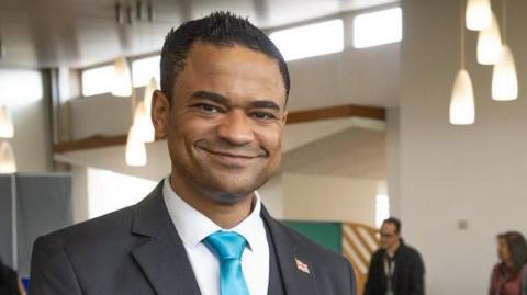 A picture of a man in a shirt and jacket with a light blue tie 