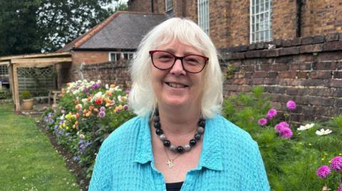A woman with white hair and a pale blue cardigan in a garden with pink flowers