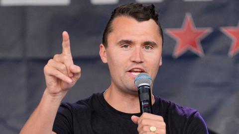 Charlie Kirk speaks into a microphone while holding up his right hand pointing his index finger. He has short dark hair and is wearing a black t-shirt.