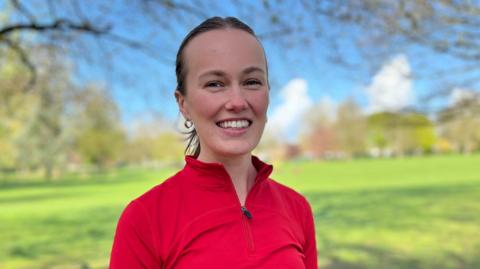 A women in a red zip up running top is smiling at the camera. She has her hair tied back and has silver hoop earrings in. She is standing in a park. 