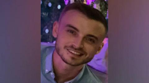 A picture of a man with short black hair smiling at the camera. He is wearing a blue button-down shirt and has black stubble