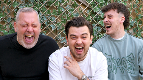 Paul, Harrison, and Travis, all laughing, in their back garden, in front of a trellis wall. Paul has short grey hair, with a beard, Harrison, dark hair and stubble and Travis, stubble, and a mullet style hair cut with the sides shaved. Paul has his eyes closed and is wearing a black top; Harrison a white T-shirt and Travis a blue top. 
