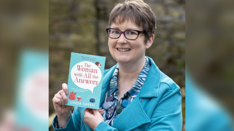 Linda Green smiling at the camera holding her book. She has short brown hair and is wearing glasses and a teal jacket over a patterned blue shirt. She is holding a book titled The Woman with All the Answers by Linda Green. The background is blurred and features an outdoor setting with a stone wall.