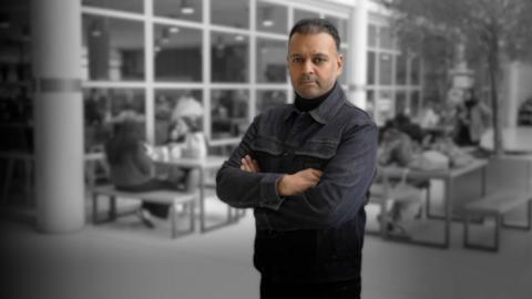 Taz Khan wears a black rollneck and dark jacket. He is looking into the camera with his arms folded across his chest