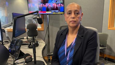 Woman with shaved head sits in a chair. She is wearing a black suit jacket over a blue and black blouse. She is sitting behind a radio microphone.