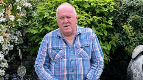 A man wearing a blue, pink and white shirt. He is sitting in a garden with his hands in his lap.