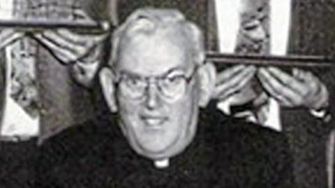 A black and white photo of the late Fr Malachy Finegan.  He has short, white hair and glasses and is wearing a black suit and a priest's collar.