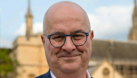 Image shows Iain Dale. He is bald and wearing black, round-rimmed glasses, and a blue suit. He is standing in front of the Palace of Westminster, which is blurred in the background.