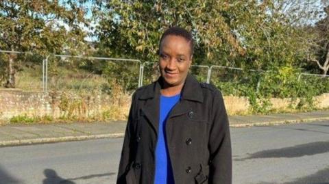 A woman with short hair in a blue top and long black jacket standing by the side of a road