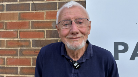 A man in a dark blue shirt, with light grey hair and glasses. He is smiling and standing in front of a brick wall.