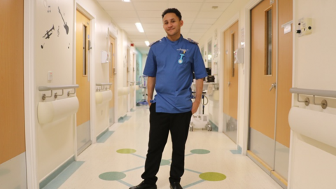 A man in a nurse's uniform stands in a hospital corridor.