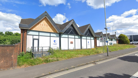 A building with several pointed gables is boarded up with overgrown grass in front of it next to a pavement and road. The sun is shining.
