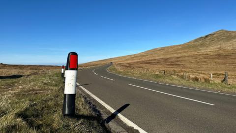 A section of the Mountain Road. There is a verge marker to the left and the peak of a hill to the right as the carriageway stretches into the distance with blue skies above.