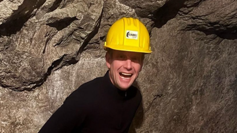 Sam Galsworthy smiles at the camera with his mouth open wearing a yellow hard hat and black jacket. There is a rockface behind him giving the impression he is inside a mine. 