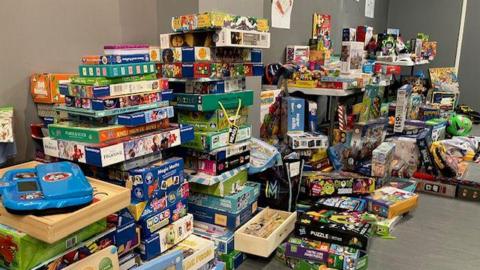 Large piles of toys in a room against a grey wall.