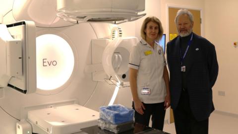 A man and female clinician standing next to a radiotherapy machine in a hospital environment. The walls and machine are white. The man has white hair and beard and is wearing a black suit, black polo neck and lanyard. The female is in a white medical jacket with short sleeves and blonde shoulder-length hair.